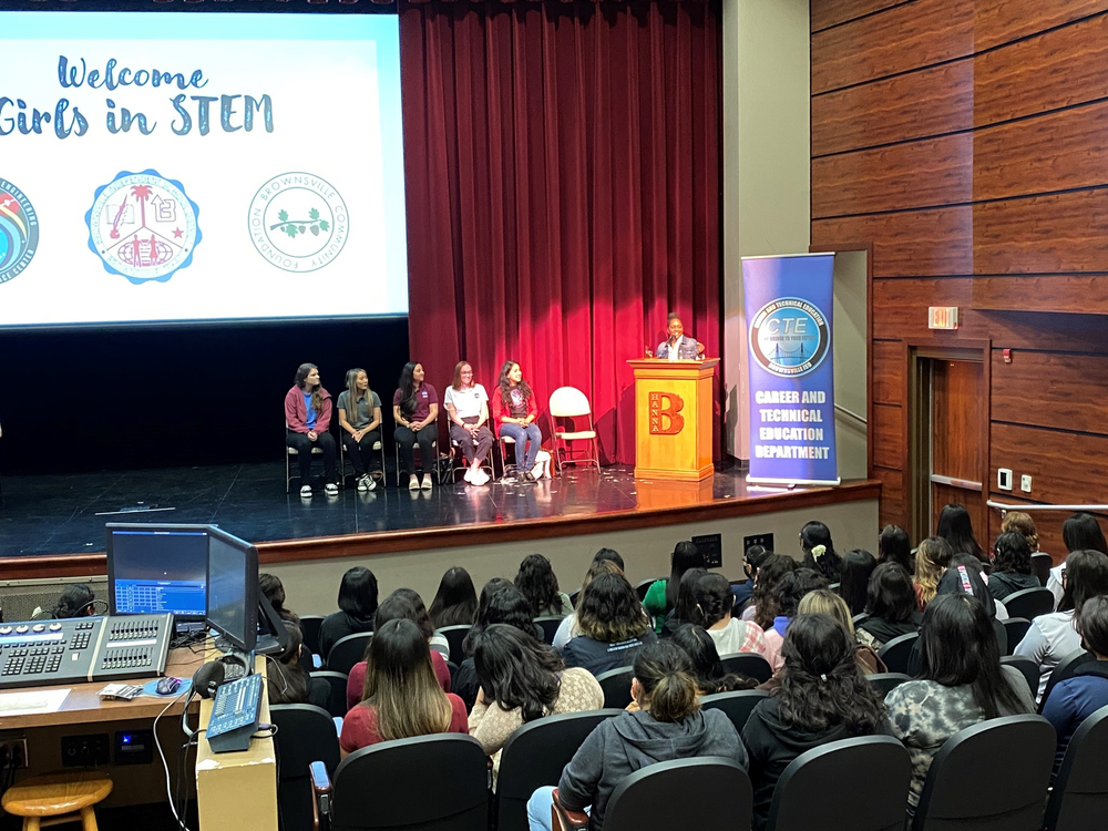 A speaker stands at a podium on a stage labeled with a "B" and a banner for the Career and Technical Education Department. On the screen behind them, text reads "Welcome Girls in STEM" along with logos. A panel of seven women is seated on the stage, facing an audience of young women.
